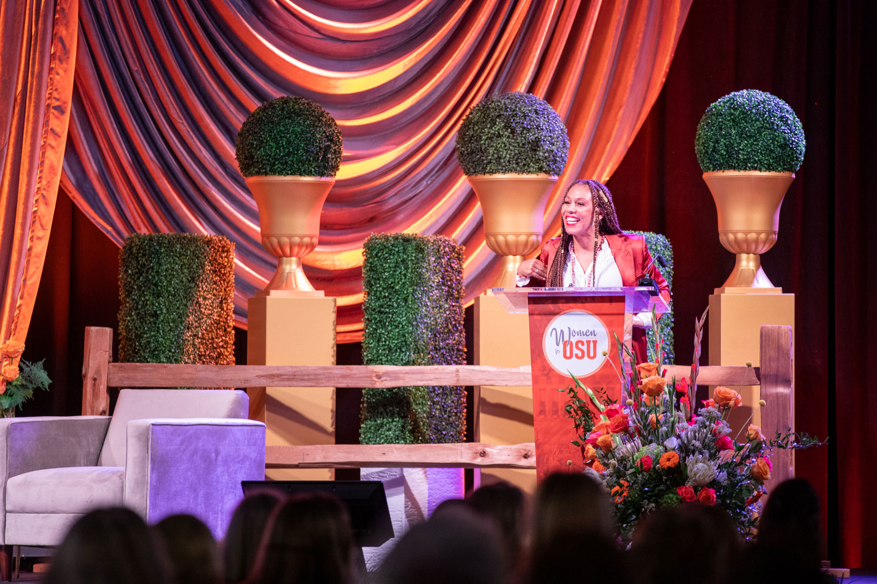 A person stands at a podium with the 'Women of OSU' logo, delivering a speech. Behind them are draped fabric and three large topiary plants in gold urns. A beige armchair is positioned to the left, and a floral arrangement sits to the right of the podium.