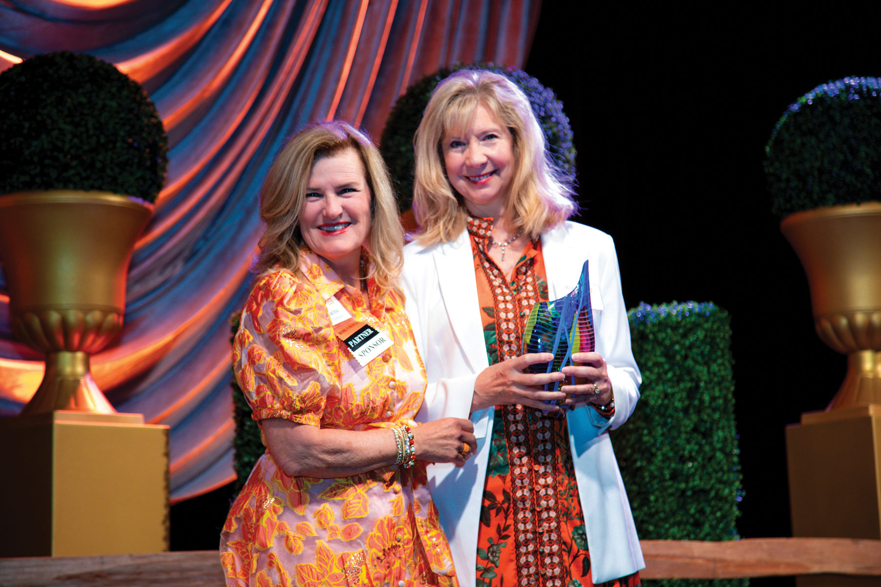 Two women stand side by side in front of a decorative backdrop with draped fabric and large potted topiary plants. The woman on the left wears an orange floral dress with a name tag, and the woman on the right wears a white blazer over a patterned dress and holds an award.