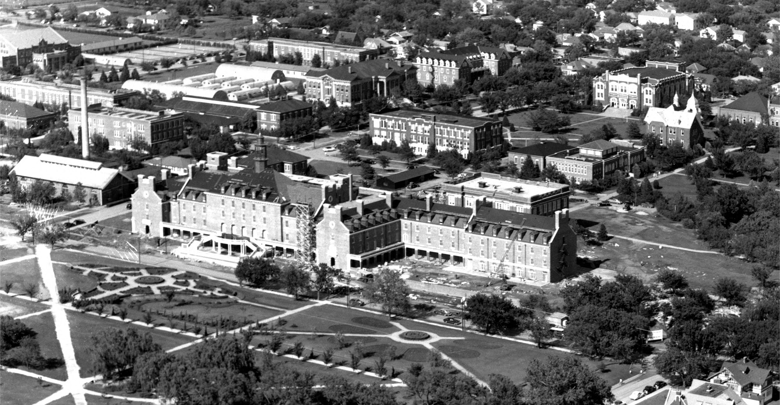 A black and white aerial photograph of a large university campus featuring multiple buildings, pathways, and green spaces. At the center is a prominent multi-story building with a U-shaped layout, surrounded by academic buildings, trees, and landscaped gardens arranged in a symmetrical design.