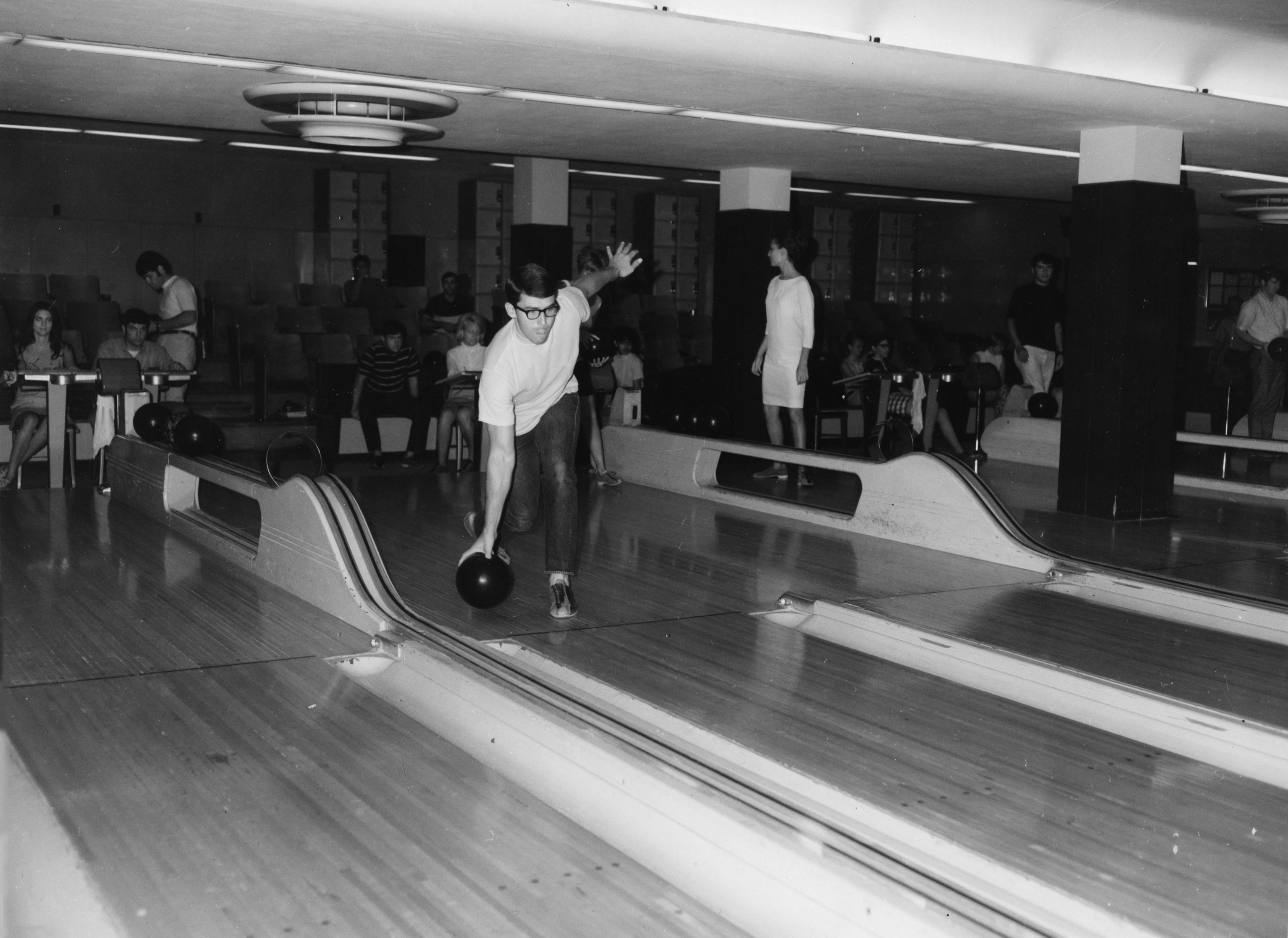 A black and white photograph of a bowling alley with multiple lanes. In the foreground, a man wearing glasses and a white shirt is in mid-action, preparing to release a bowling ball.