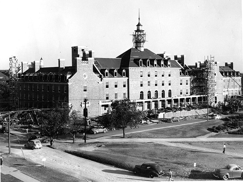 A black and white photograph of a large, multi-story brick building with a central tower featuring an ornate structure. The roof includes multiple chimneys and dormer windows. 