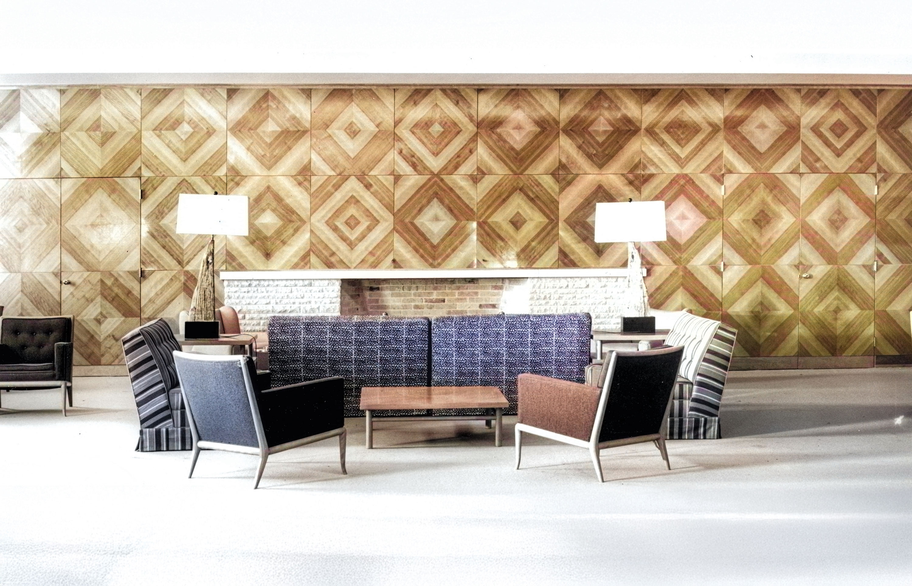 A modern lounge area featuring a geometric wooden wall with diamond-shaped patterns in various shades of brown. The seating includes armchairs in black, brown, and striped upholstery, two white floor lamps, and a central low wooden coffee table. 