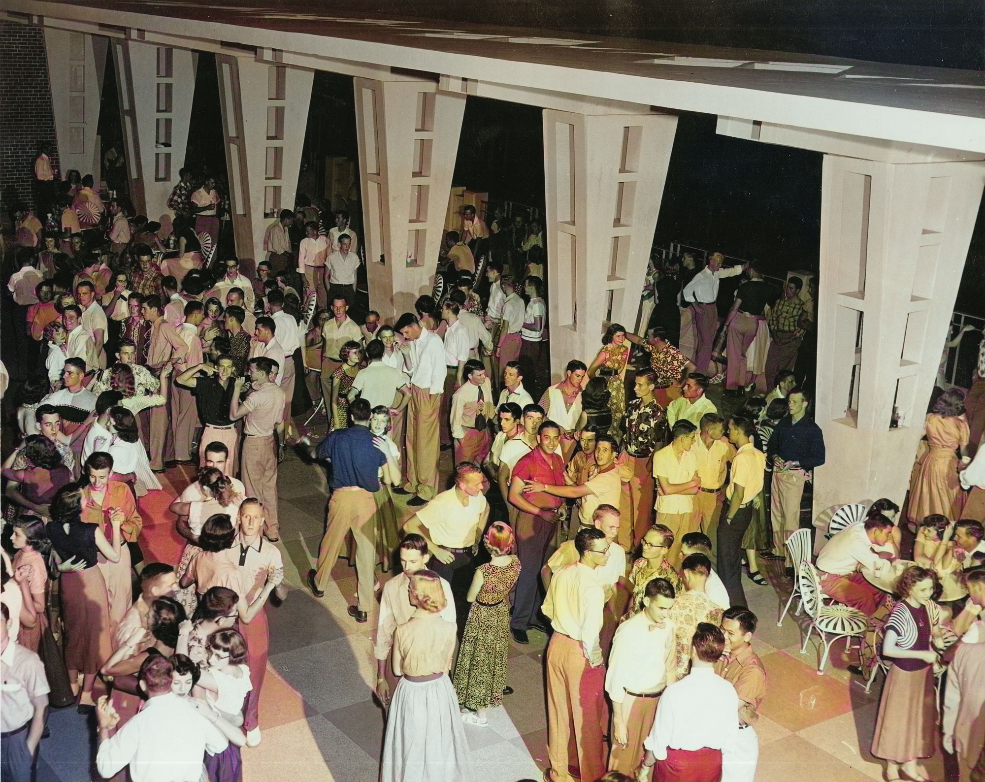 A large group of people gather in a spacious, well-lit area with tall white columns. Dressed in mid-20th century attire, men wear slacks and button-up shirts, and women wear skirts and blouses.