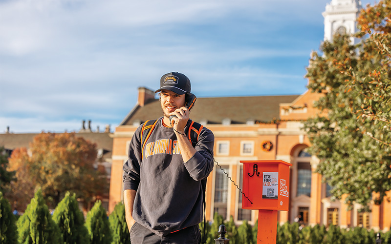 Person wearing an Oklahoma State sweatshirt and a cap, standing outdoors near an orange emergency phone station with campus buildings and trees in the background.