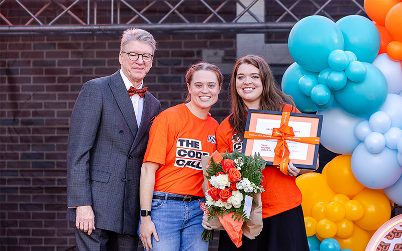 Three people standing in front of a colorful balloon arrangement, with two wearing bright orange shirts holding a bouquet of flowers and a framed certificate, and one dressed in a dark suit with a bow tie.