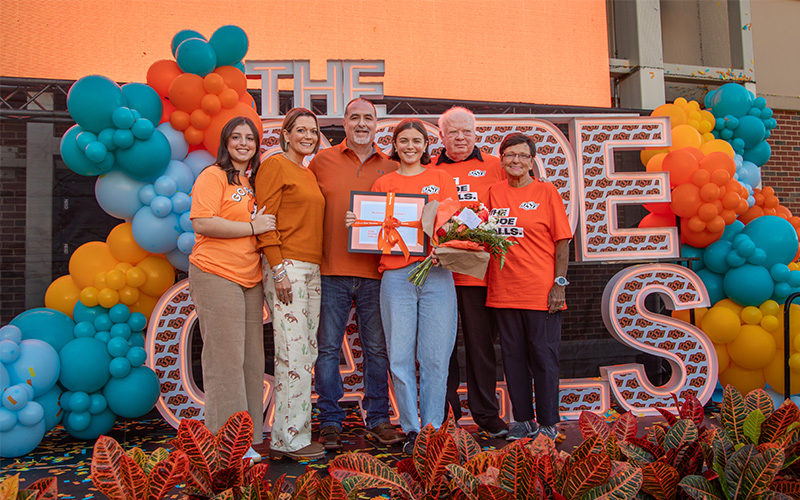 Group of six people standing on a stage in front of large letters spelling ‘THE CODE CALLS,’ surrounded by colorful balloon arrangements in orange, blue, and yellow. One person holds a bouquet of flowers and two framed certificates.