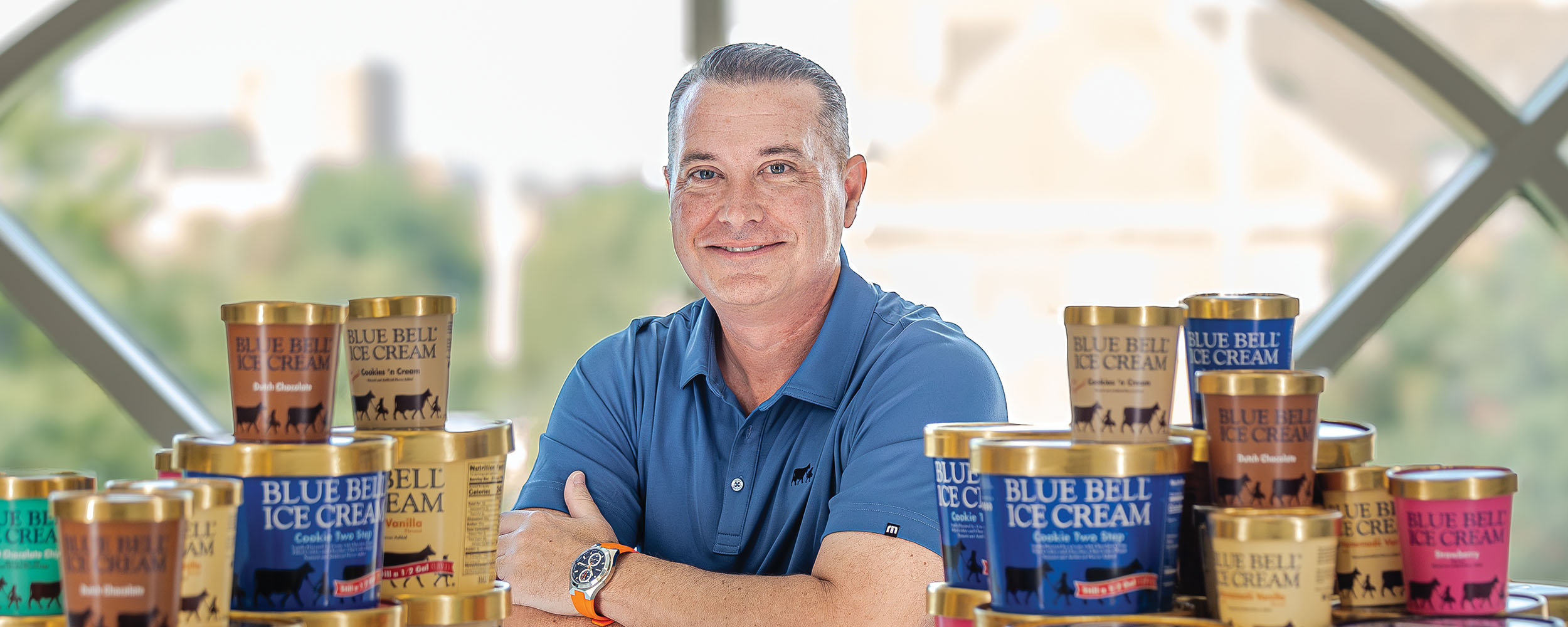 A person sits at a table with arms folded, surrounded by multiple containers of Blue Bell Ice Cream arranged in stacks and rows. The ice cream containers display a variety of flavors in different colored cartons. A large arched window in the background shows a softly blurred outdoor campus setting.