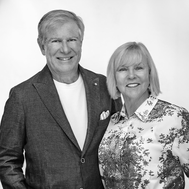 Two people standing side by side against a plain background, one wearing a dark textured jacket over a light shirt with a pocket square, and the other wearing a patterned floral blouse.