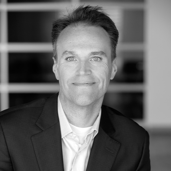 Person wearing a dark suit jacket over a light-colored dress shirt, seated indoors with a blurred background featuring horizontal window panels.