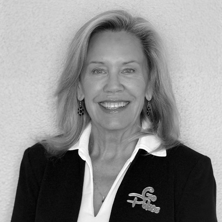 Person wearing a dark blazer with a white collared shirt and a decorative brooch that reads ‘Go Pokes,’ standing against a light textured wall.