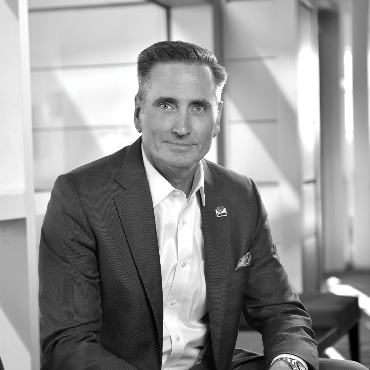 Person wearing a dark suit jacket over a white dress shirt with a pocket square and a lapel pin, seated indoors with light streaming through modern panel walls in the background.