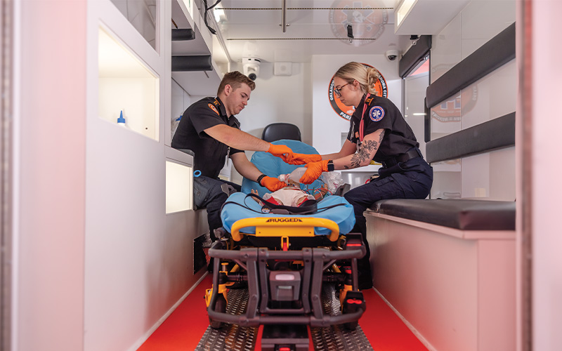 Workforce Development Two emergency medical personnel inside an ambulance attending to a patient on a stretcher, wearing dark uniforms and orange gloves, with medical equipment visible in the background.