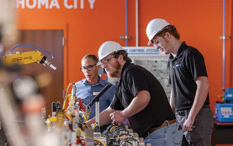 Three people wearing safety helmets working with industrial automation equipment in a lab setting, surrounded by wires, control panels, and a robotic arm, with an orange wall in the background displaying partial text.