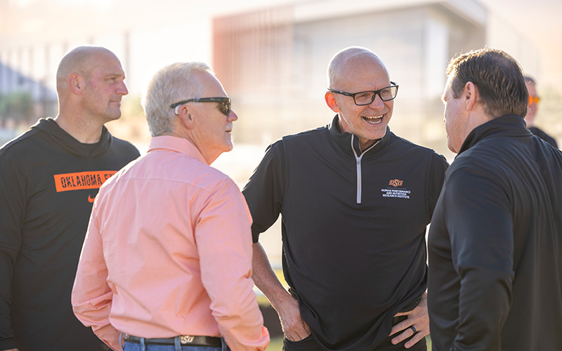 Four people stand in a small group outdoors, talking during what appears to be an event. They wear a mix of casual and branded OSU apparel, including a quarter‑zip pullover and an orange button‑down shirt. A modern building and other attendees are visible in the softly lit background.
