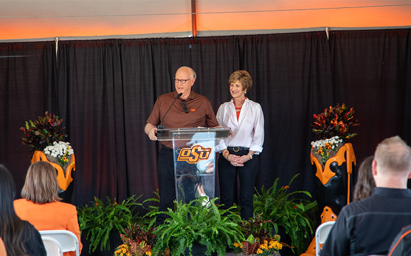 A speaker stands behind a clear podium with the OSU logo during an indoor event, with another person standing beside them. The stage is decorated with black curtains, orange-and-black floral arrangements, and green plants at the front. Audience members seated in rows face the stage.
