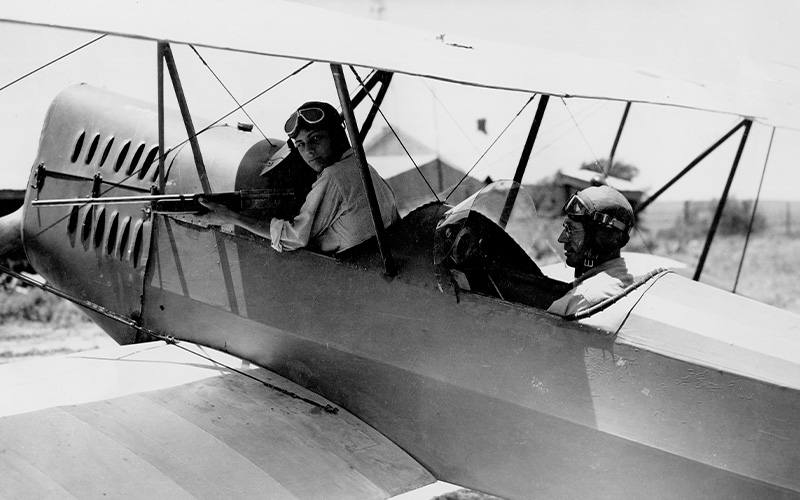 Flying Farmers Two people sit in an open‑cockpit biplane on the ground. The person in the front cockpit faces forward wearing flight goggles and a cap, while the person in the rear cockpit appears to be operating or inspecting a mounted machine gun. The aircraft’s fabric-covered wings, struts, and fuselage are clearly visible, with an open landscape and a few distant structures in the background.