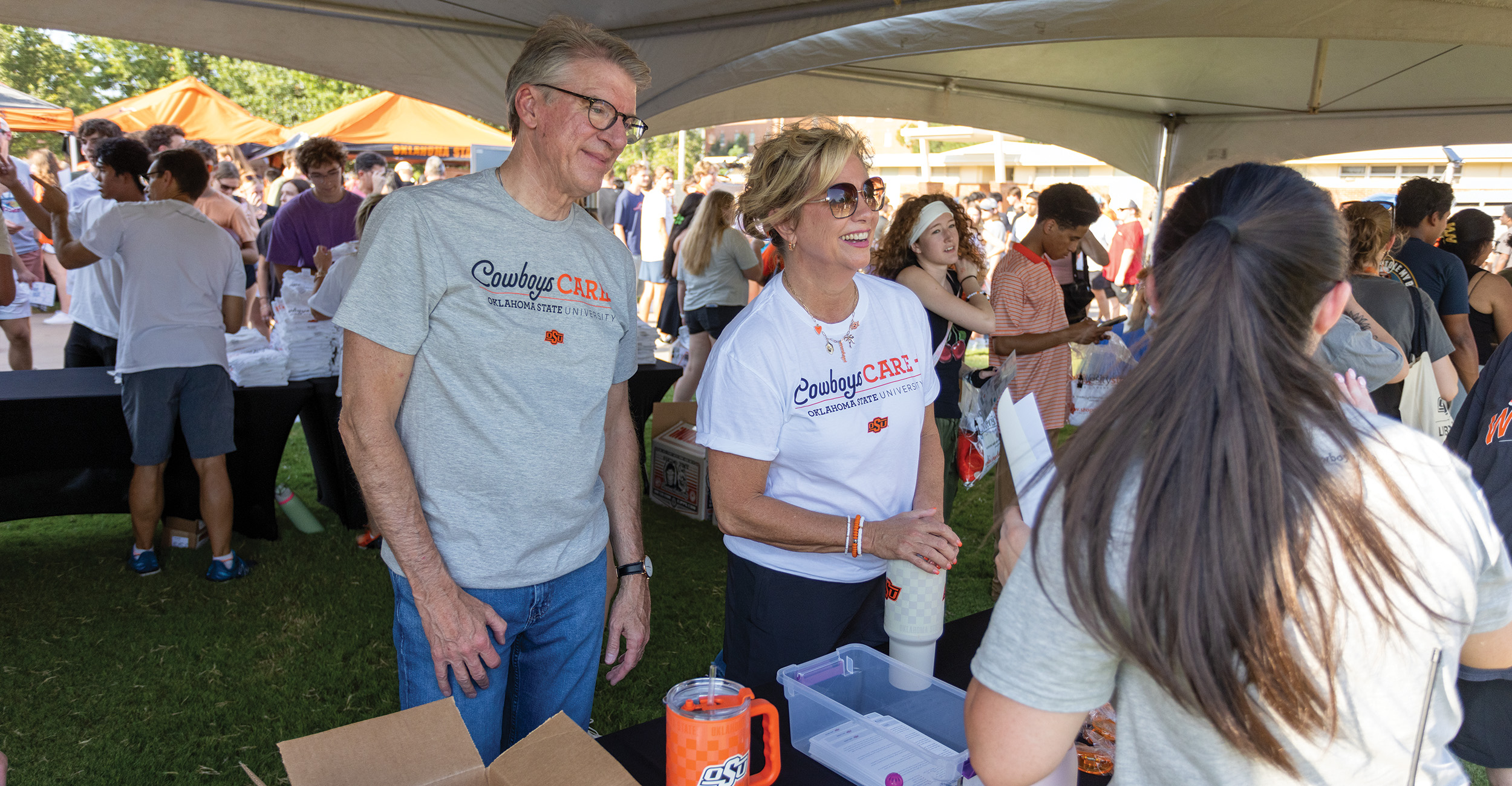 Two people wearing gray and white ‘Cowboys Care Oklahoma State University’ T-shirts standing under a canopy at an outdoor event, with tables, boxes, and a crowd of attendees in the background.