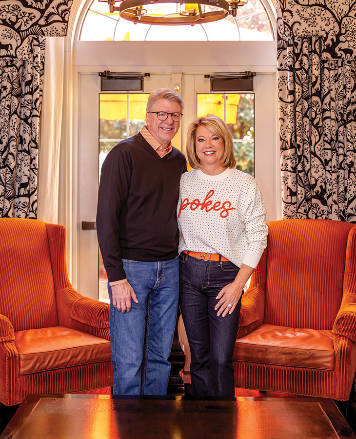 A man and a woman are standing side by side in a well-decorated room. The man is wearing a black sweater and blue jeans, while the woman is wearing a white sweater with the word 'pokes' written on it in orange letters and dark jeans. They are standing between two orange armchairs with patterned curtains behind them and a window that lets in natural light.
