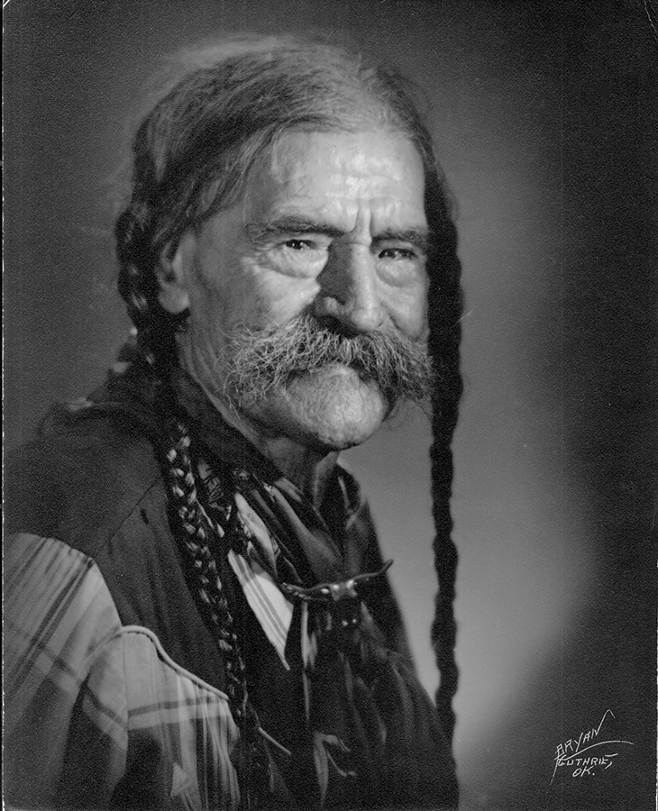 An elderly man with long braided hair, wearing a plaid shirt and a bolo tie. The photograph is in black and white.