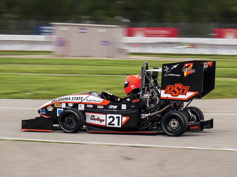 A race car with the number 21, representing Oklahoma State University, is captured in motion on a racetrack. The car features prominent branding with "OSU" and "Cowboys" logos.