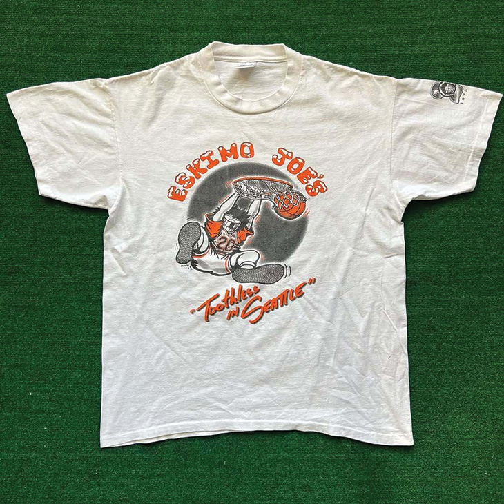 A white T-shirt with a graphic design on the front. The design features a cartoon character of a man wearing an orange and white sports jersey duniking a basketball. The text above the character reads 'Eskimo Joe's' in red letters, and below it says 'Stillwater, OK' in smaller red letters. The T-shirt is laid flat on a green surface."