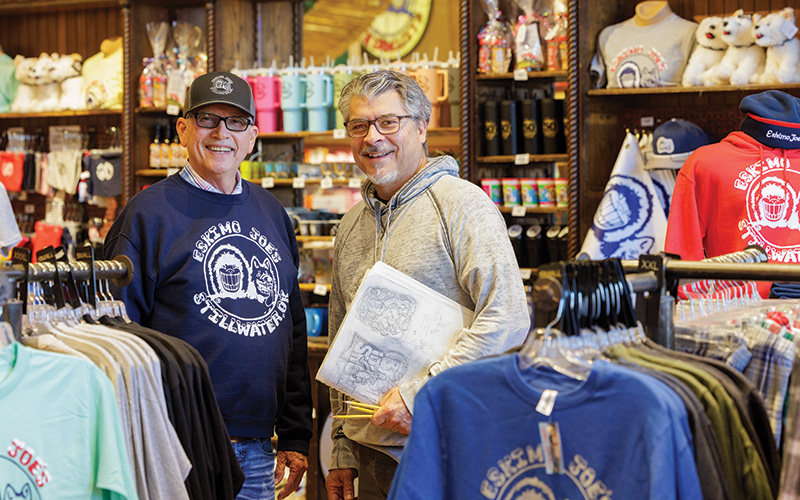 Two men are standing in a clothing store. The man on the left is wearing a navy blue sweatshirt and a black cap, while the man on the right is wearing a gray hoodie and holding a white bag with a design on it. The store has various clothing items displayed, including t-shirts, sweatshirts, and hats with 'Eskimo Joe's Stillwater' logos. There are also stuffed animals and other merchandise visible in the background.
