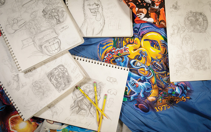 Several sketchbooks with pencil drawings are spread out on a colorful t-shirt. The sketches include various cartoonish characters, some of which are smiling or laughing. Three yellow pencils are placed on top of one of the sketchbooks. The t-shirt features a vibrant design with animated characters and the text 'A Fun House 1975-2015.' This image showcases the creative process behind character design and illustration, highlighting both rough sketches and a finished product on the t-shirt.