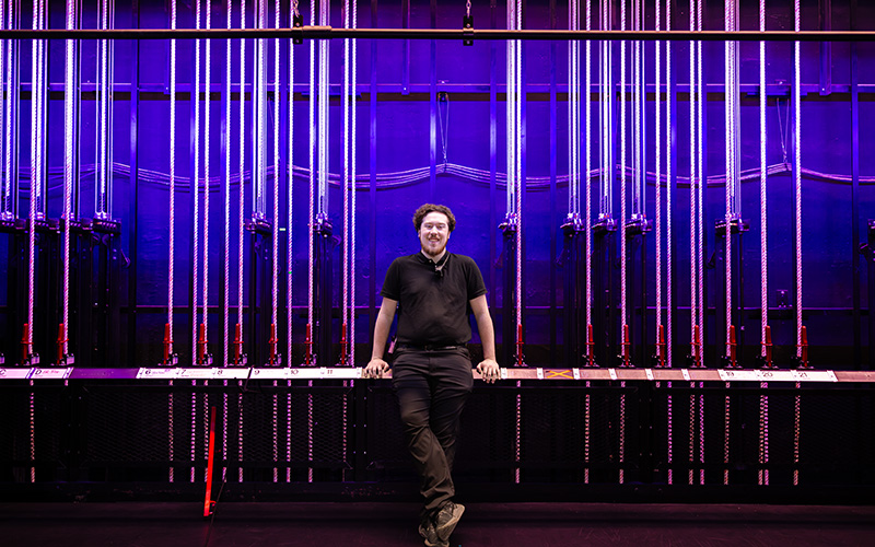 A man is standing in front of a wall with multiple vertical light tubes emitting purple and blue light. He is wearing a black shirt and pants, with arms resting on the railing behind them and legs crossed at the ankles. The background features an array of illuminated tubes creating a visually striking effect