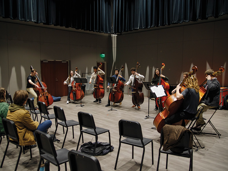 A group of eight people, appearing to be a mix of men and women, are playing double basses in a music rehearsal room. They are arranged in a semicircle with music stands in front of them. There are empty chairs scattered around the room, and a few people are seated, observing the rehearsal. The room has high ceilings with dark curtains and acoustic panels on the walls.