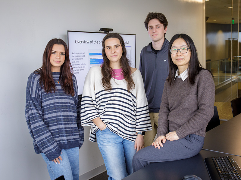  Four people are standing and sitting in a modern office setting. They are dressed casually in sweaters and jeans. Behind them is a large screen displaying a presentation titled "Overview of the project portal." 