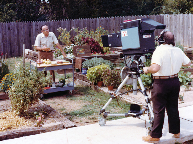 A television production scene in an outdoor garden. A man stands behind a cart displaying vegetables and gardening tools, speaking as part of a segment. Raised garden beds with lush plants surround him. A cameraman operates a large vintage studio camera labeled 'OETA,' capturing the scene.