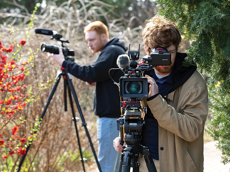 Two people are operating professional video cameras outdoors. The person in the foreground is wearing a brown jacket and looking through the viewfinder of a Sony camera mounted on a tripod, with various accessories attached to the camera. The person in the background, whose face is blurred, is also using a camera on a tripod and appears to be focusing on something off-frame. There are red flowers and greenery visible in the background, indicating that this scene takes place in a natural setting.