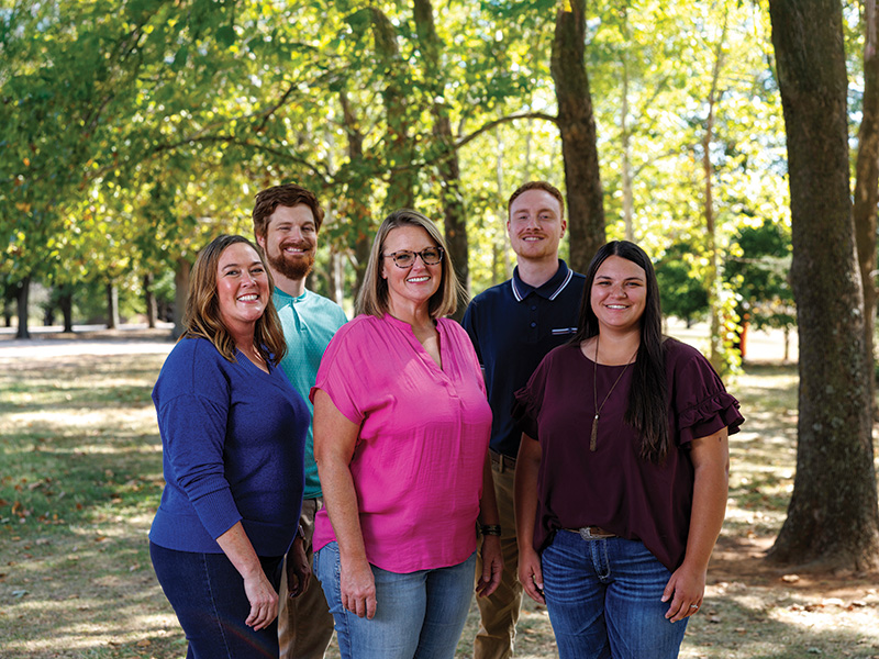 A group of five people standing outdoors in a park-like setting with trees and greenery in the background. The individuals are dressed casually, with three women and two men.