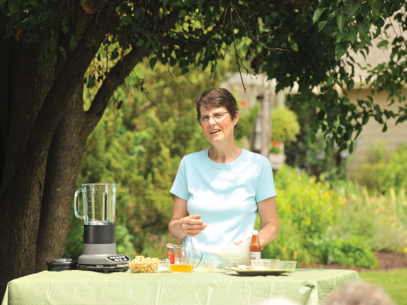 A woman in a light blue shirt stands behind a green tablecloth-covered table outdoors, under a tree. On the table are various items including a blender, small bowls containing ingredients like nuts and liquid, and a bottle of sauce. The background features greenery and part of a house.