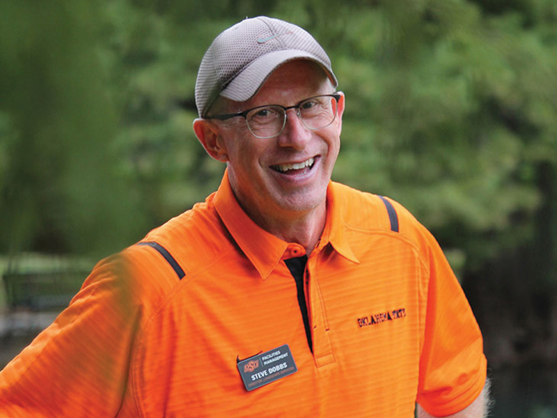 A man wearing an orange shirt with black accents and a gray cap. The shirt has a name tag that reads 'OSU' and 'Steve Dobbs.' The background is blurred greenery.