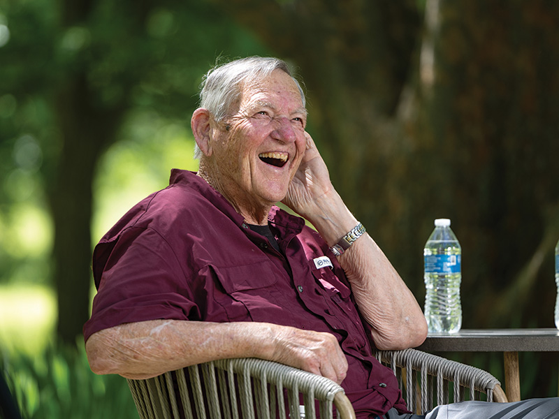 An elderly man with gray hair sits outdoors in a wicker chair, smiling and laughing with his hand to his face. He wears a maroon shirt and a wristwatch. A bottle of water sits on the table next to him, and green trees and sunlight fill the background.