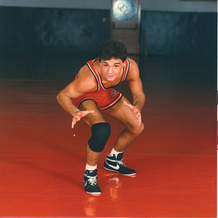 A man wearing an orange wrestling singlet, black knee pads, and black Nike shoes is in a wrestling stance on an orange mat.