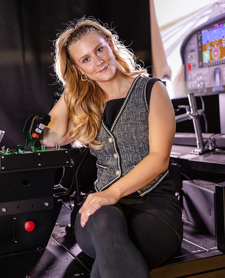 A woman with long, wavy blonde hair is sitting in a flight simulator cockpit. She is wearing a sleeveless black top and a grey vest with white trim. Her right hand is on the control stick, and she appears to be engaged in operating the simulator. The cockpit features various controls and displays, including a screen showing flight information. The setting suggests an environment related to aviation training or simulation.