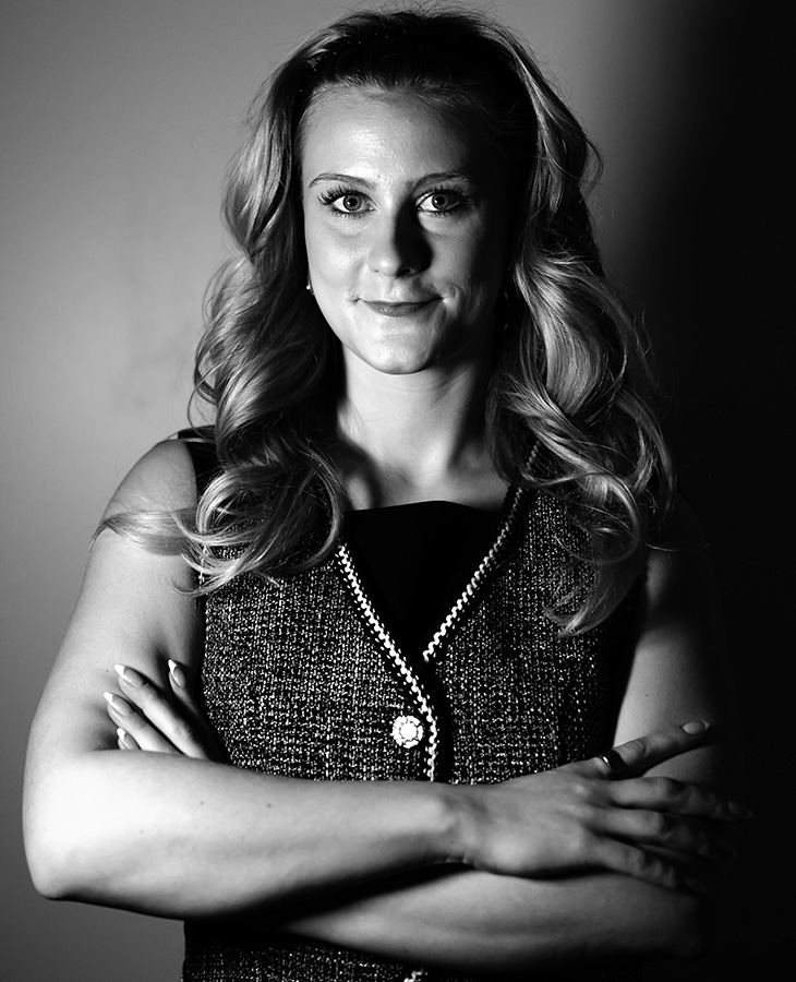 A black and white photograph of a woman with long, wavy hair, wearing a sleeveless dress with a textured pattern and a decorative button at the center. Her arms are crossed in front of their chest.