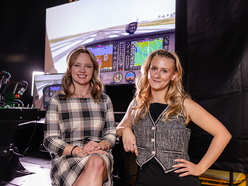 Two women are sitting in front of a flight simulator setup. The woman on the left is wearing a plaid dress, and the woman on the right is wearing a black sleeveless top with a gray vest. The flight simulator screen behind them displays various aircraft controls and navigation information.