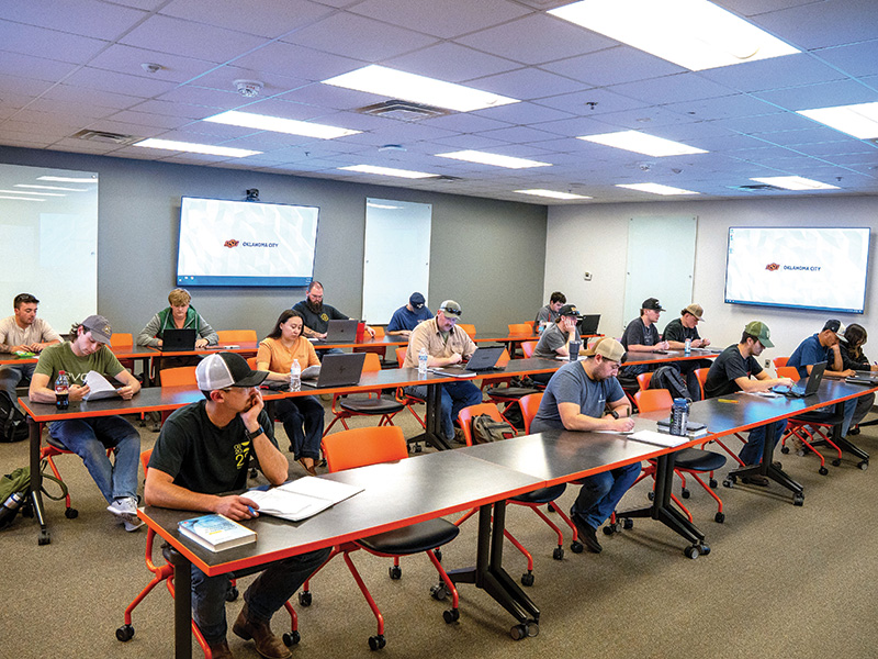 A classroom with several people sitting at desks, working on laptops or writing in notebooks. The room has orange chairs and long tables arranged in rows. There are two large screens at the front of the room. 