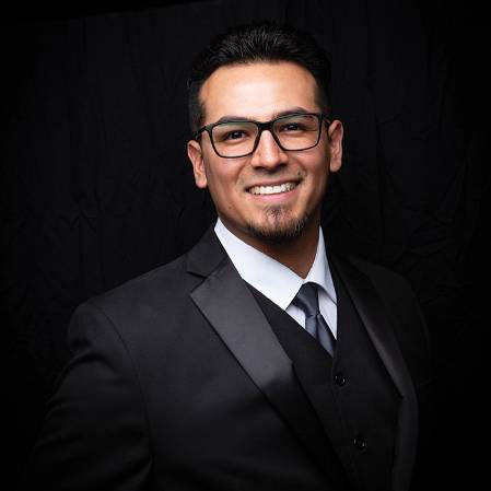 A person who appears to be a man is wearing a black suit, white dress shirt, and a gray tie, standing against a dark background.