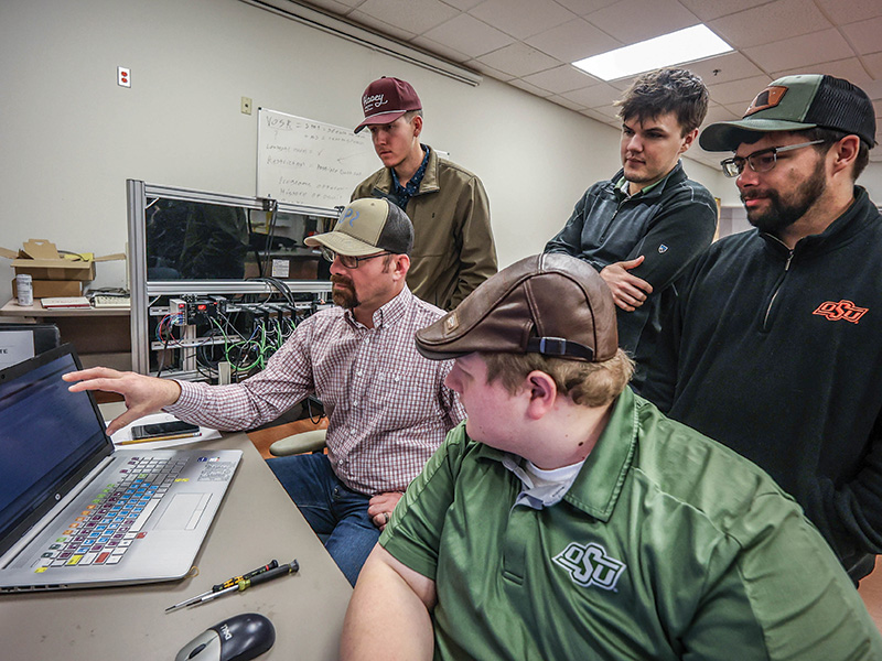 A group of five people are gathered around a laptop in an office setting. One person is seated and pointing at the laptop screen, while the others stand around observing. The individuals are wearing casual clothing, including baseball caps and shirts with "OSU" logos. In the background, there is a whiteboard with some writing on it and various electronic equipment on shelves.
