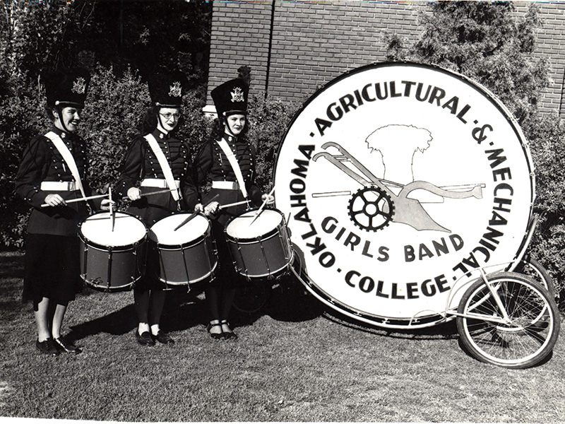 A black and white photograph shows three members of the Oklahoma Agricultural & Mechanical College Girls Band standing outdoors in uniform, each holding a snare drum. They are positioned next to a large bass drum on a wheeled stand, which displays the band's name and logo featuring an agricultural emblem. The background includes foliage and part of a brick building.