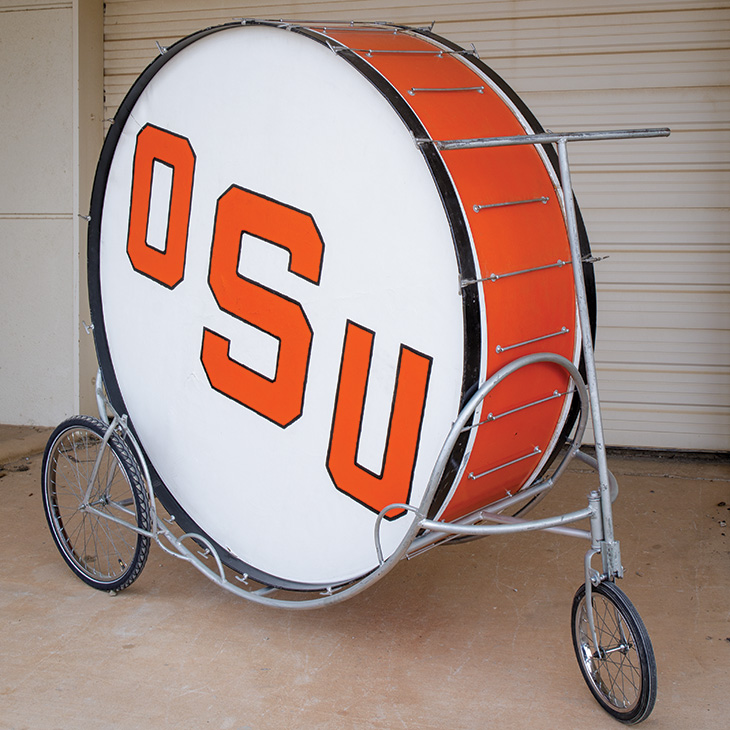 A large bass drum with the letters 'OSU' in bold orange font on a white background. The drum is mounted on a metal frame with three wheels for mobility. The sides of the drum are orange, and it is positioned in front of a closed garage door.