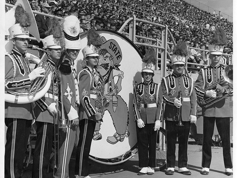 A black and white photograph of a marching band standing in front of a large bass drum with the words 'STATE' written on it. The drum features an illustration of a cartoonish figure wearing a hat and holding a baton. The band members are dressed in elaborate uniforms with tall hats adorned with plumes, epaulets, and decorative sashes. They are holding various brass instruments, including tubas and trumpets. In the background, there is a large crowd seated in stadium bleachers.