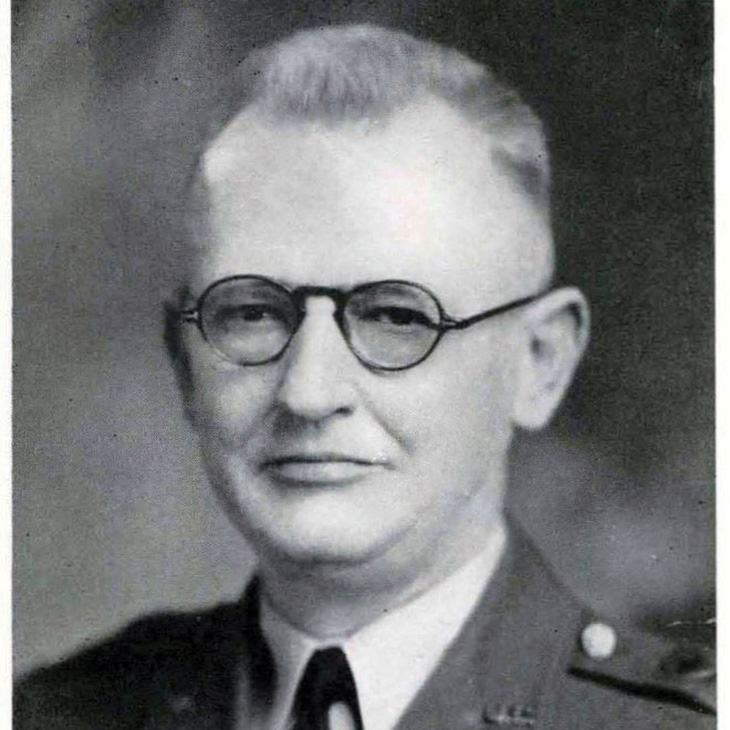"A black and white photograph of a man in military uniform. The uniform includes a jacket with epaulets and a tie. The background is plain and dark, focusing attention on the subject's attire.