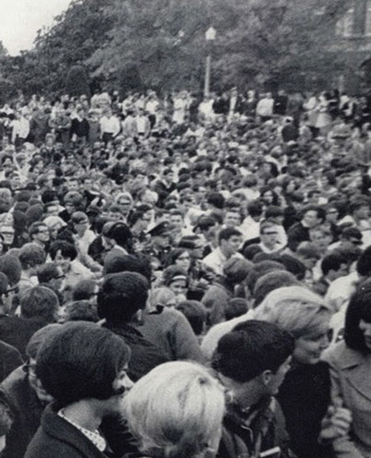 A large crowd of people gathers on a campus lawn in this historic black-and-white photograph, with attendees filling the foreground and stretching into the distance.