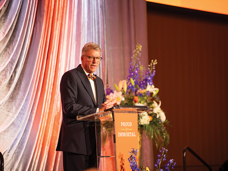 A speaker stands at a podium during a formal Oklahoma State University event, delivering remarks in front of decorative drapery and large floral arrangements.