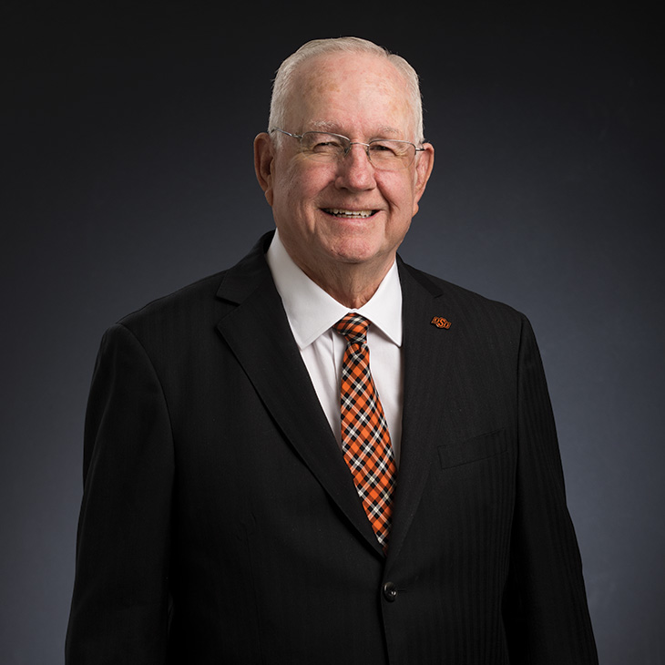 A person in a dark suit and patterned tie poses for a professional headshot against a dark studio background.